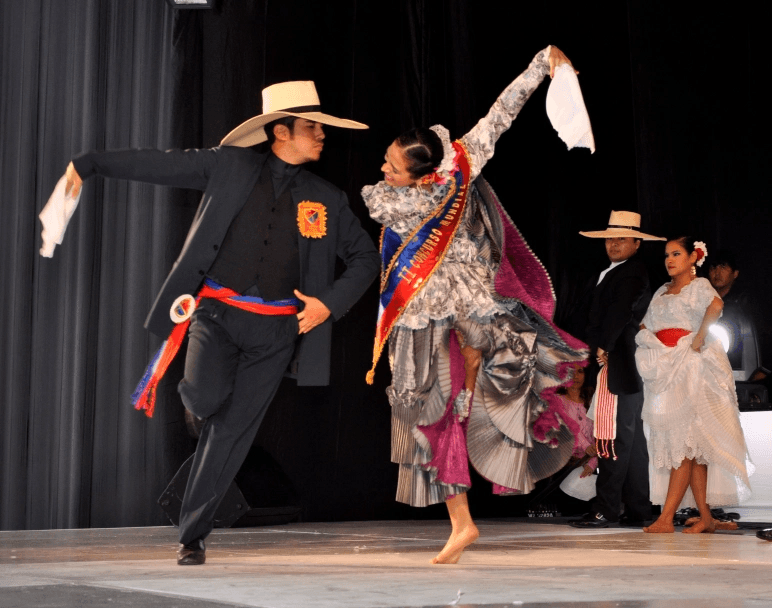Bailarín con traje negro y sombrero alza el pañuelo junto a su pareja en un ensayo sobre el escenario