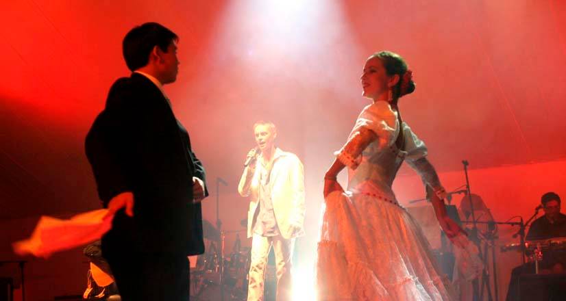 Pareja de marinera sobre un escenario con luces rojas y humo mientras un cantante actúa al fondo