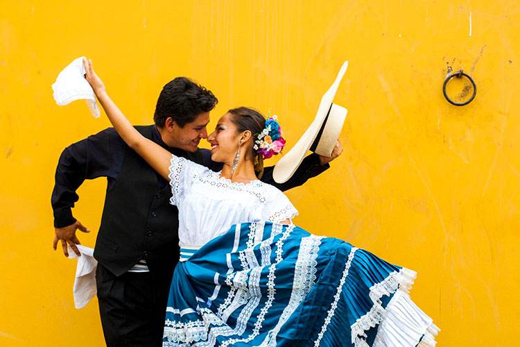 Pareja de marinera norteña frente a una pared amarilla: ella con falda azul y blanca y él con sombrero de paja y pañuelo