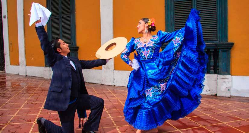Pareja de marinera en una calle colonial: él arrodillado con el pañuelo en alto y ella con vestido azul bordado