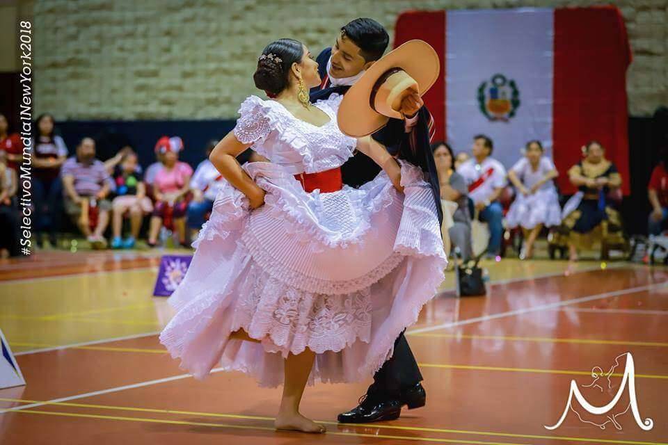 Pareja bailando marinera en un selectivo con la bandera del Perú de fondo, ella con vestido blanco de encaje